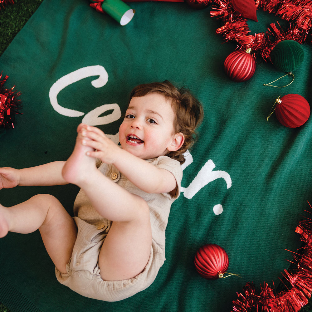 
                  
                    Load image into Gallery viewer, Child lying on a green blanket with Christmas decorations.
                  
                