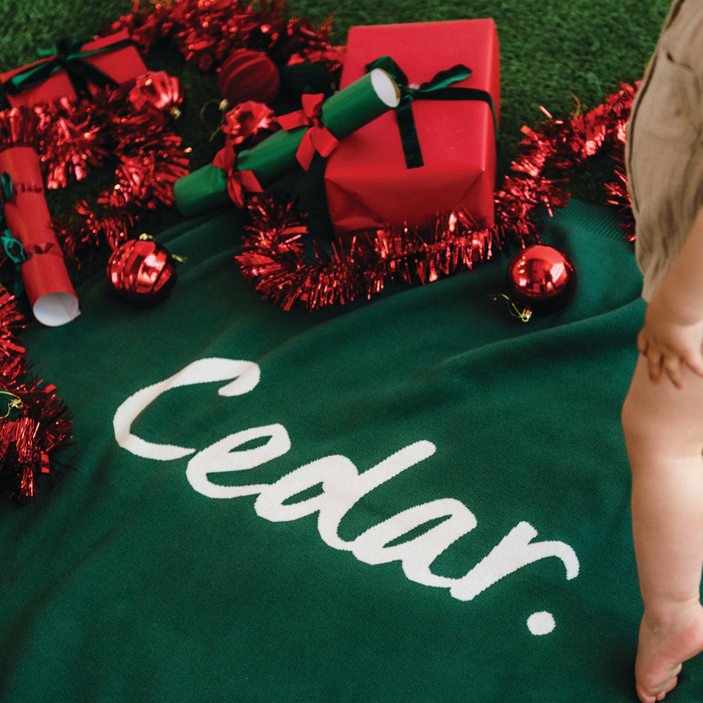 Green blanket with the name 'Cedar.' knitted into it, red gift, and Christmas decorations on a grassy background