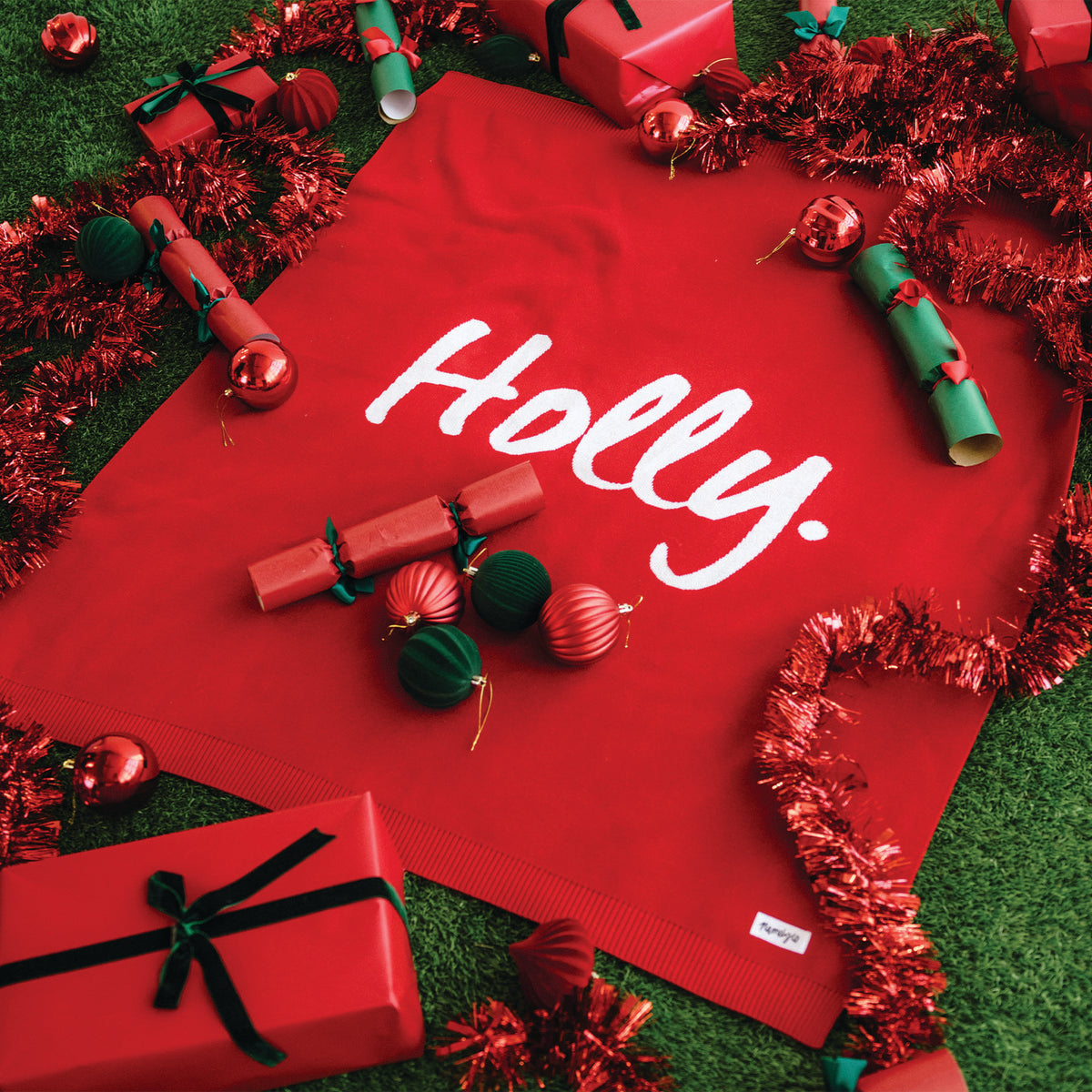 Red blanket with 'Holly' text surrounded by Christmas decorations on a green surface