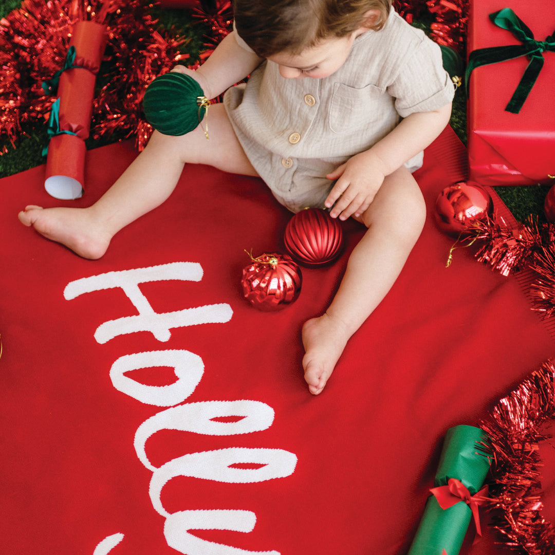 Child sitting on a red blanket with Christmas decorations and the word &